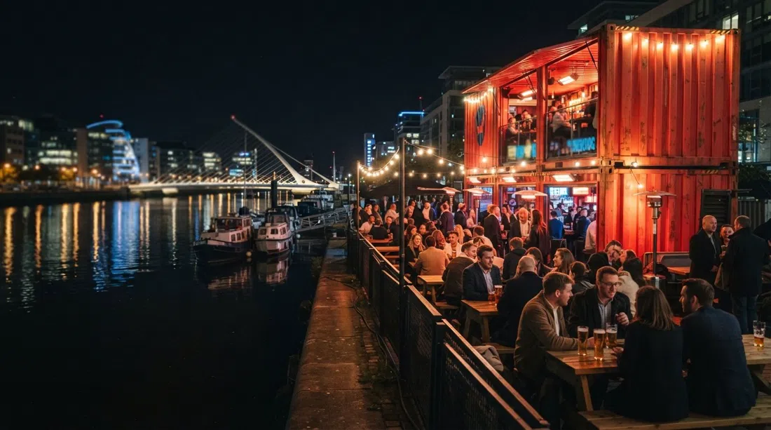 밤의 Capital Dock에 있는 BrewDog Dublin, 강 옆에서 빛나는 빨간색 컨테이너 바로, 난방 테라스와 물에 비친 도시 불빛